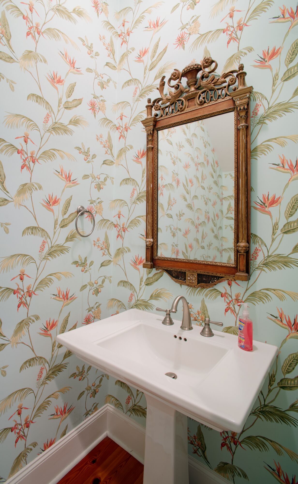 Un baño con papel tapiz floral, un lavamanos blanco y un espejo con marco de madera.