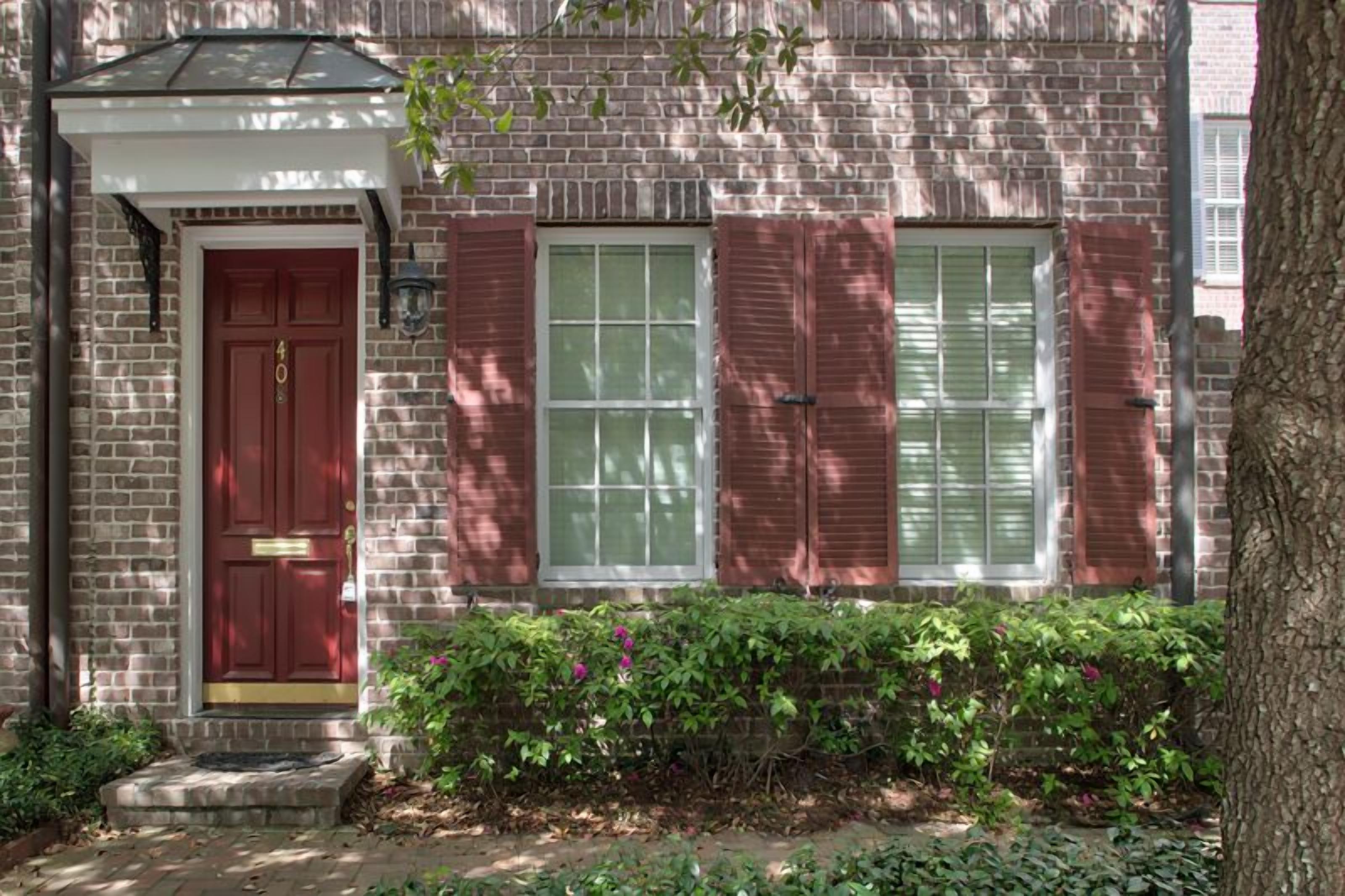 Una casa de ladrillo con puerta roja, dos ventanas con contraventanas y un pequeño porche.