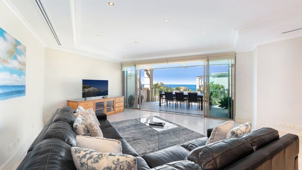 A modern living room with a leather sofa, a glass coffee table, and a TV on a wooden cabinet. There is a sliding glass door leading to an outdoor patio with a view of the ocean. - Unit 46  - 3 Bed Ocean View (TERRIGAL)