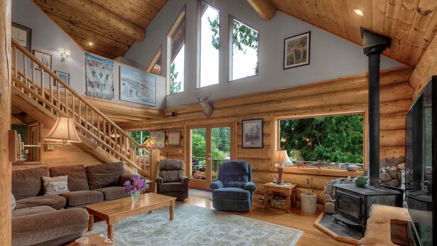 A spacious living room with a wooden staircase, a fireplace, and large windows. - Bonny Sky Lodge- private riverfront log cabin w/ mountain view, hot tub, generator backup! (Gold Bar)