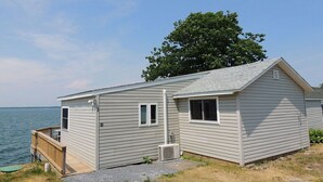 Ein eingeschossiges Blockhaus mit einer Terrasse, einem kleinen Anbau und einem großen Baum im Hintergrund.
