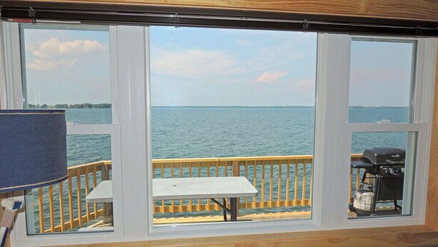 Ein Zimmer mit großem Fenster mit Blick auf ein Gewässer, eine Holzterrasse mit Tisch und Grill sowie eine Couch im Vordergrund.