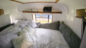 Un intérieur chaleureux avec un lit, des oreillers, une étagère ornée d’objets décoratifs et une fenêtre donnant sur un paysage agréable.