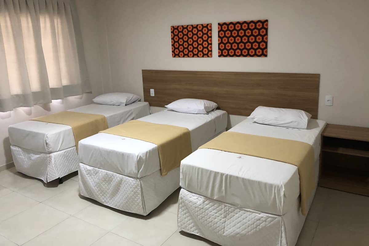 Quarto de hotel com três camas individuais, cada uma com colchão branco e manta bege. Duas obras de arte fixadas na parede exibem um padrão geométrico.