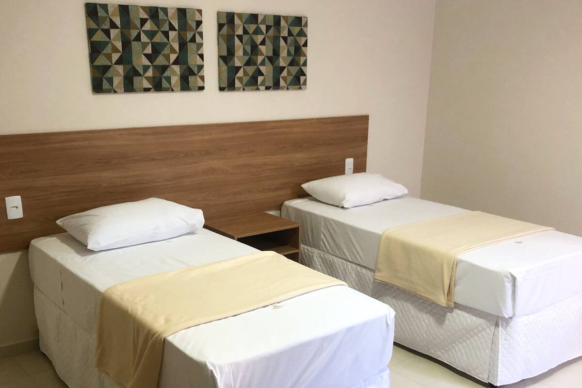Duas camas de solteiro com roupa de cama branca e cabeceira de madeira, uma pequena mesa de cabeceira e duas obras de arte geométricas penduradas na parede.