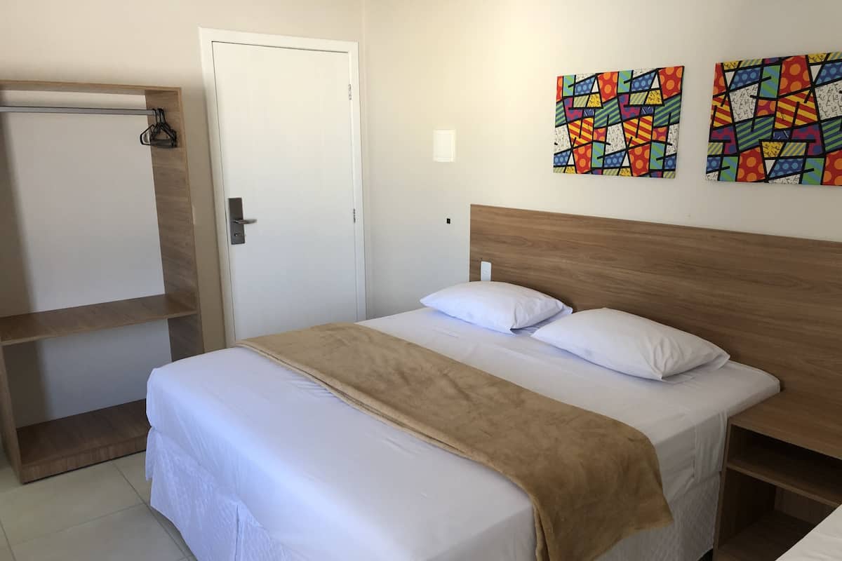Cama bem arrumada com manta marrom, cabeceira de madeira e duas pinturas abstratas coloridas na parede.