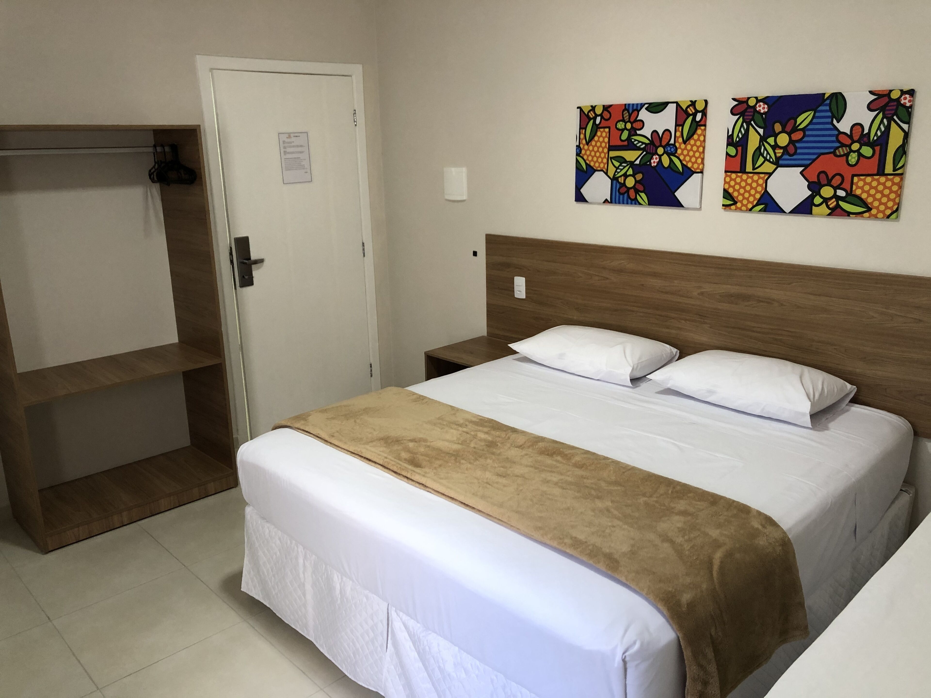 Une chambre d’hôtel avec un lit, une tête de lit en bois, une table de chevet, une porte blanche et deux œuvres d’art colorées accrochées au mur.