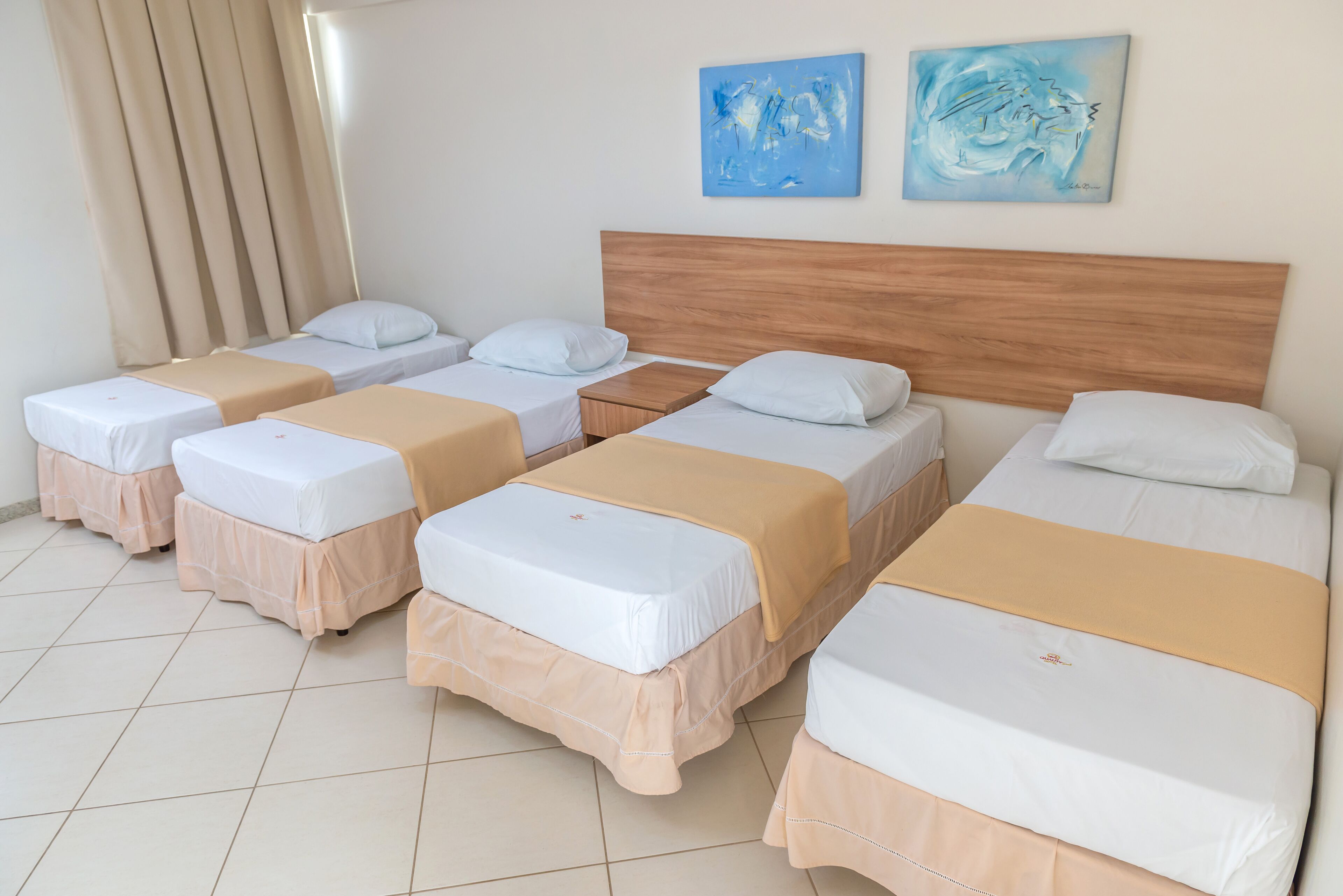 A hotel room with four single beds, beige and white bedding, a wooden headboard, and two abstract paintings on the wall.