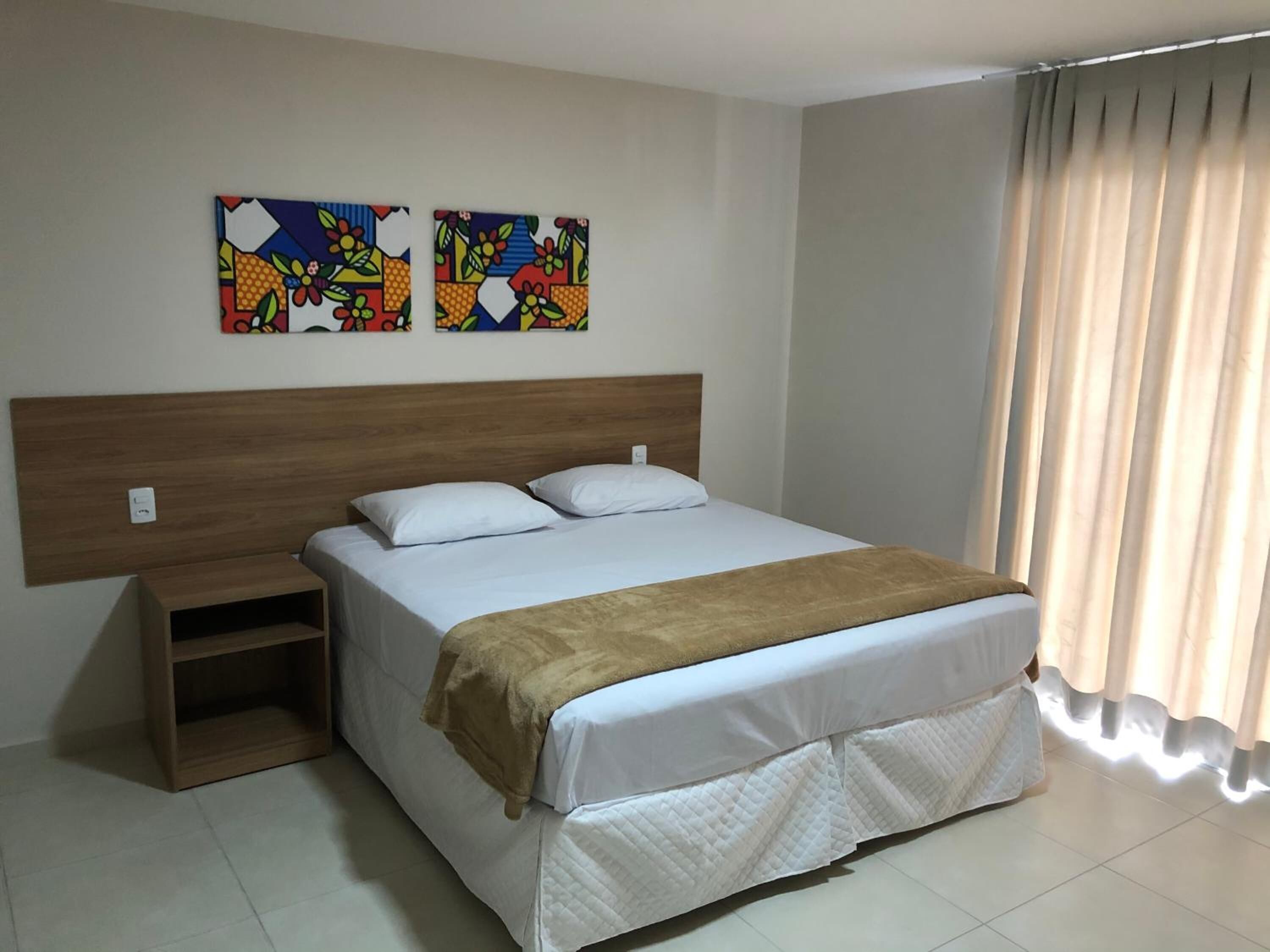 Quarto de hotel com cabeceira de madeira, cama com roupa de cama branca e manta marrom, duas pinturas coloridas na parede e uma janela com cortinas.