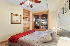 Une chambre à coucher avec un lit, un ventilateur de plafond, une fenêtre donnant sur des arbres, une bibliothèque et un miroir.