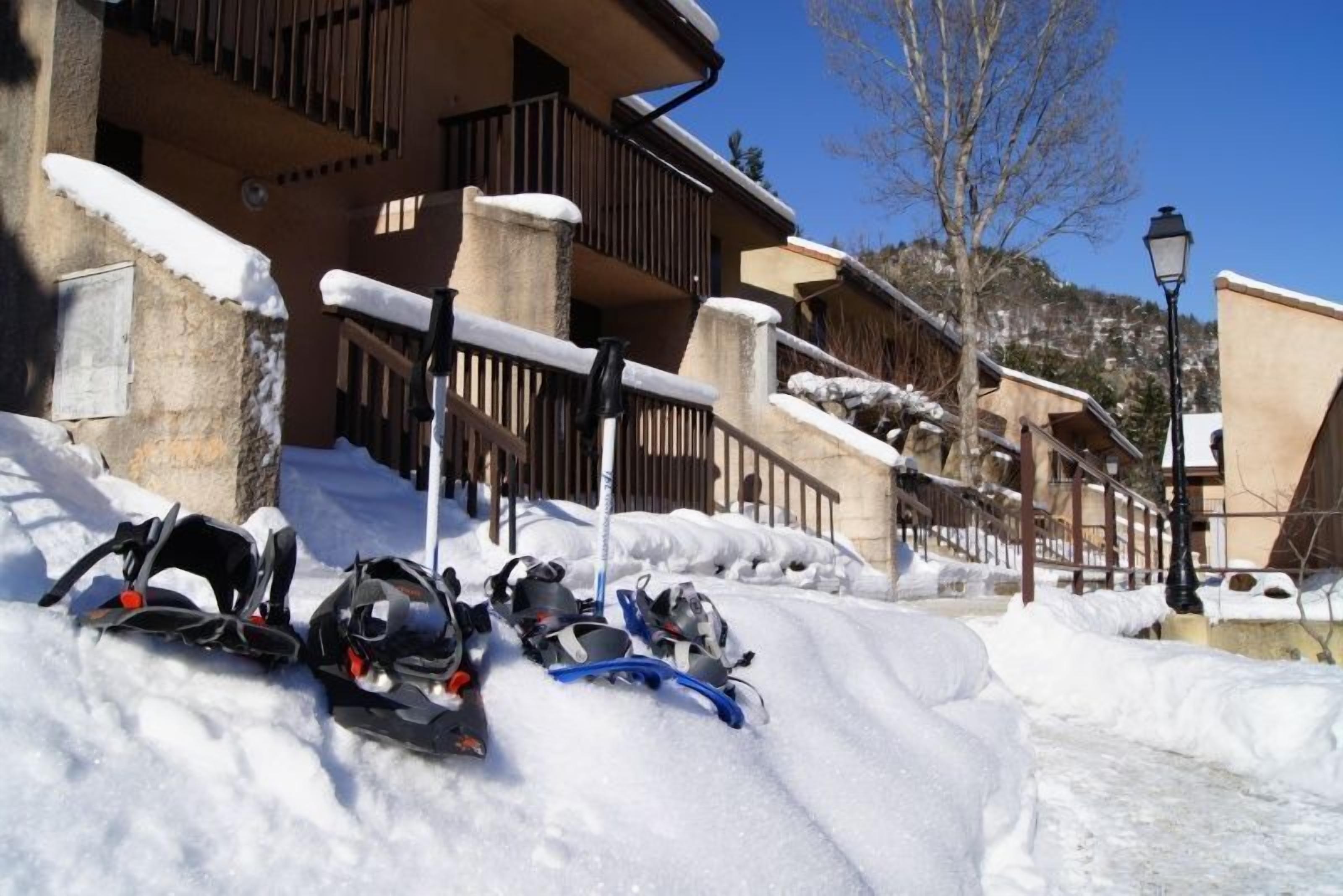 A snow-covered street with snowshoes and skis in the foreground, a building with balconies and snow on the roof, and a clear blue sky in the background.