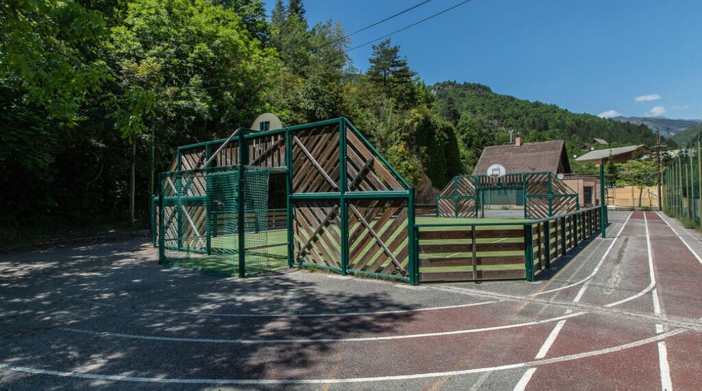 Un terrain de sport clôturé, équipé d’un panier de basket, entouré d’arbres et avec une montagne en arrière-plan.