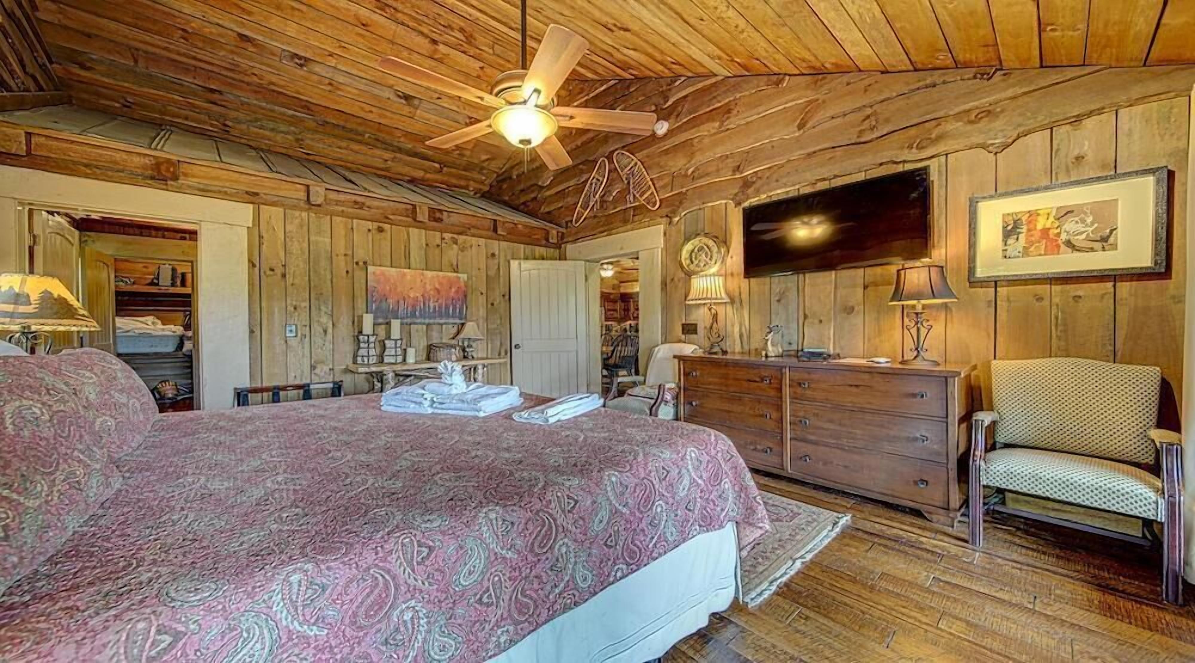 A bedroom with a large bed, wooden ceiling, a ceiling fan, a dresser, a chair, and a painting on the wall.