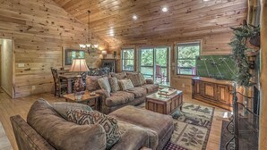 Interior acogedor de una cabaña con techo de madera, un conjunto de sofás, una mesa de centro, chimenea y un ventanal con vistas a la vegetación.