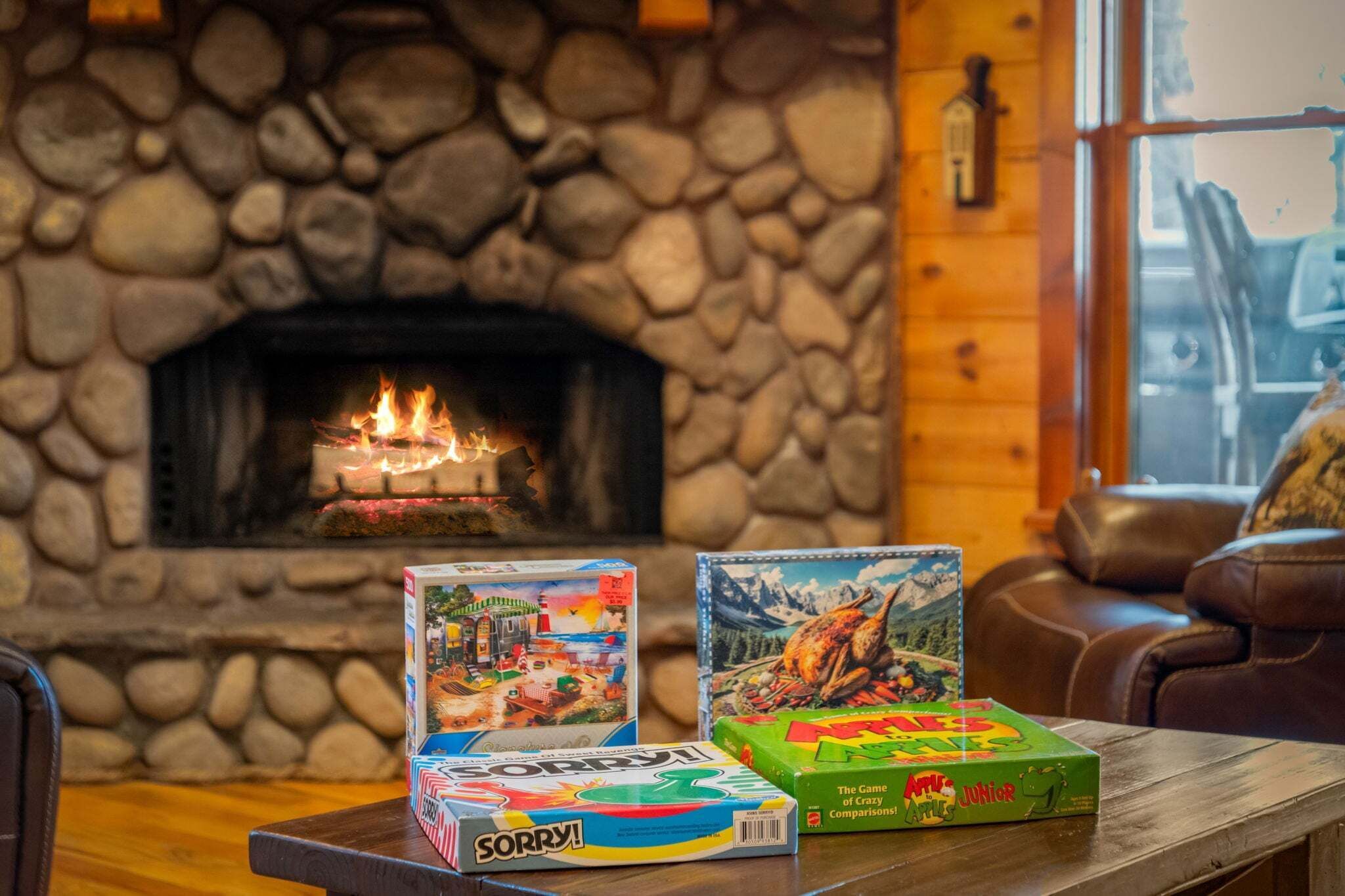 A fireplace with a lit fire, a stone wall, a wooden table with board games, and a leather couch.