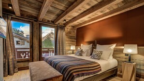 Een slaapkamer met een houten plafond, een groot bed met een gestreept dekbed, twee nachtkastjes met lampen, en uitzicht op bergen door een raam.