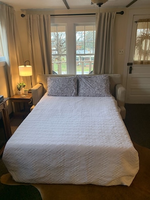 Una cama bien hecha con un cojín estampado, una lámpara de noche y vistas al exterior a través de una ventana.