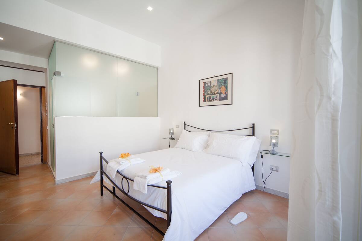 A hotel room with a bed, a nightstand, a wall-mounted lamp, a painting, and a door.