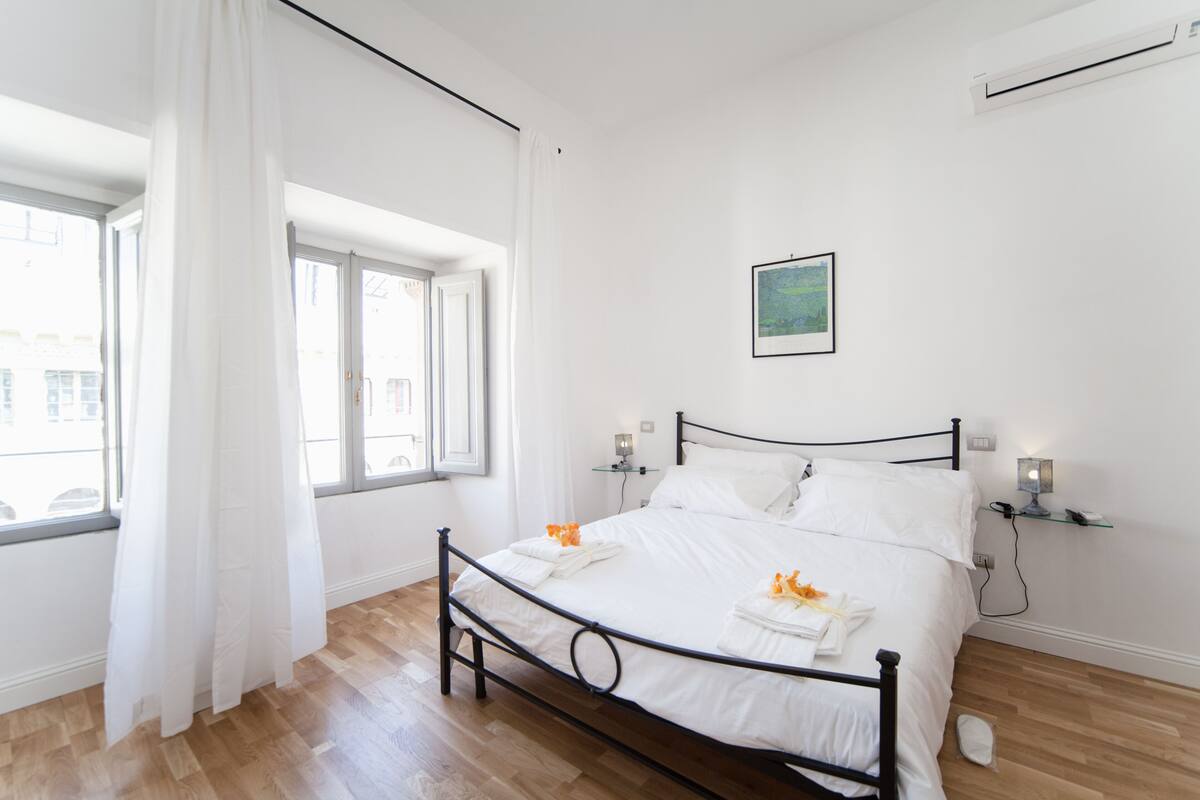 A neatly made bed with white linens and towels, a bedside table with a lamp, and a window with curtains.