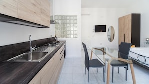 A modern kitchen with a dark countertop, stainless steel sink, and wooden cabinets. There is a dining area with a glass table and black chairs. A TV is mounted on the wall, and a mirror is visible. - Eugenia e Donato (Naples)