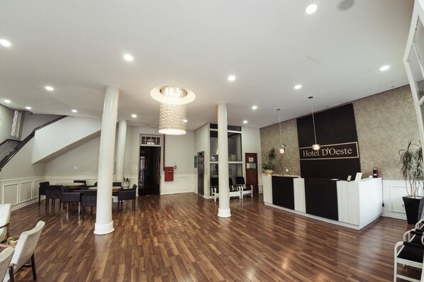 A hotel reception area with a wooden floor, white columns, a reception desk, and a staircase. - Hotel D'Oeste (Poços de Caldas)