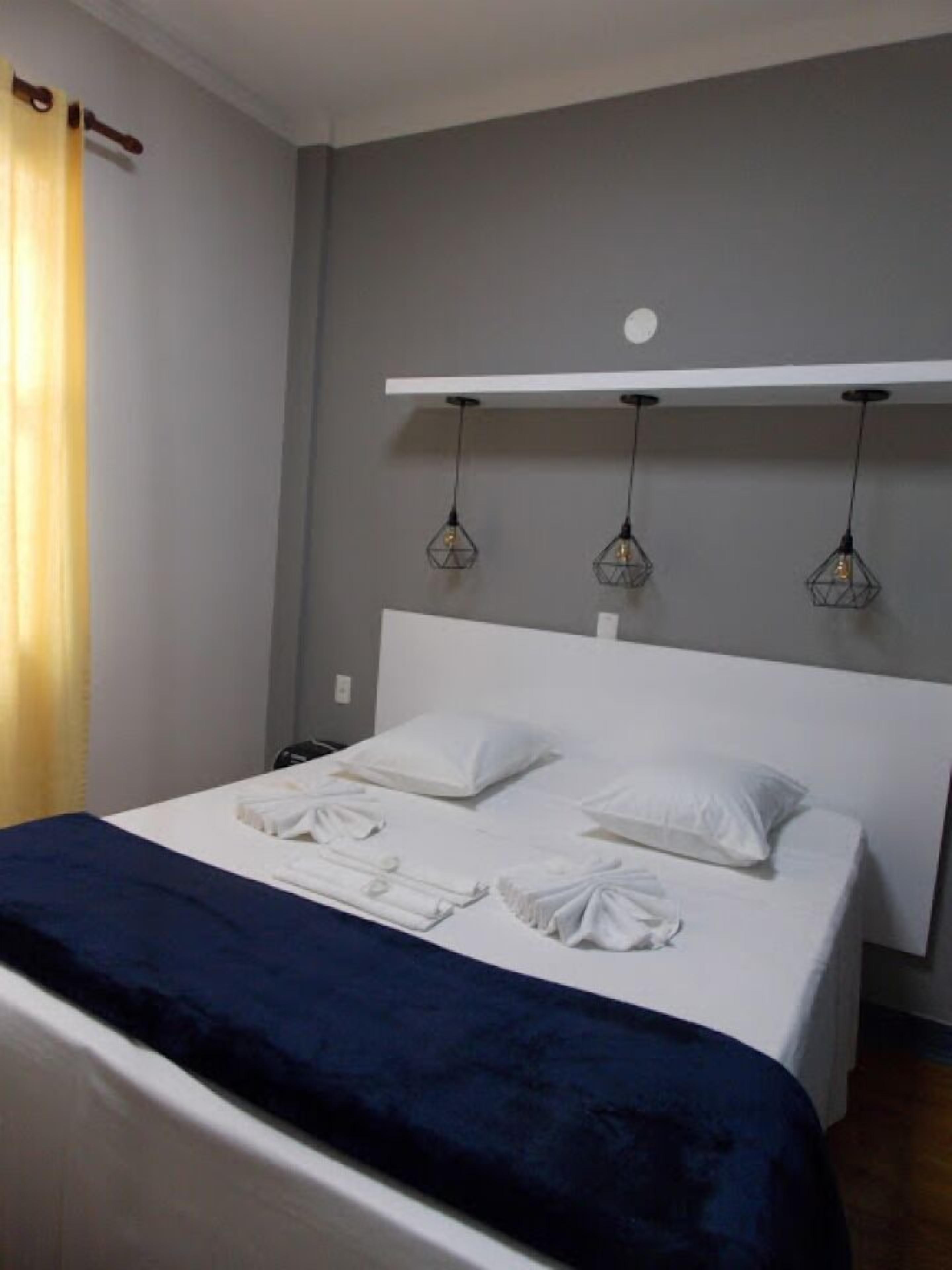 Un lit bien fait, avec du linge de lit blanc et une couverture bleu foncé, une tête de lit blanche et trois lampes suspendues dont les ampoules sont apparentes.