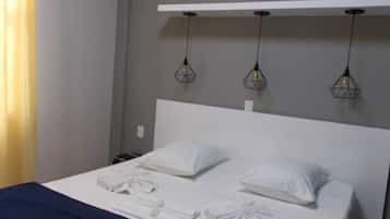 Un lit bien fait, avec du linge de lit blanc et une couverture bleu foncé, une tête de lit blanche et trois lampes suspendues dont les ampoules sont apparentes.