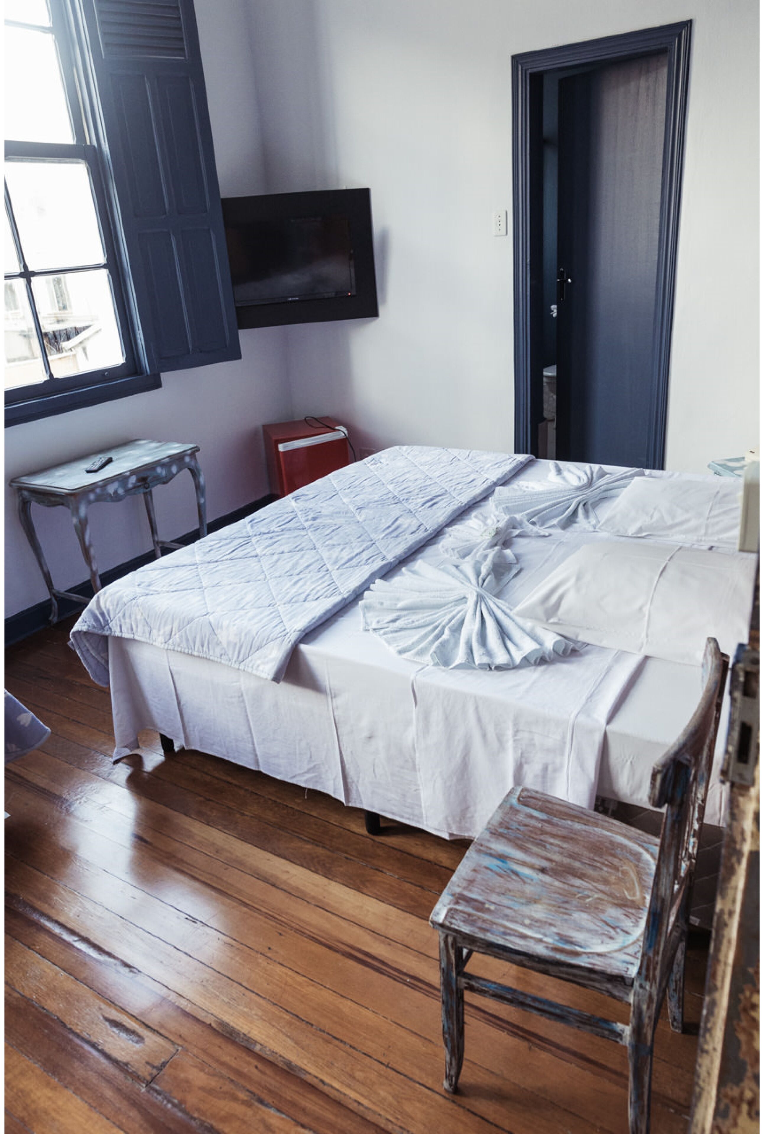Un dormitorio con cama, televisor montado en la pared, ventana y piso de madera.
