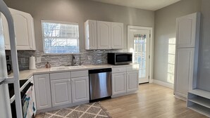 Una cocina moderna con armarios blancos, lavavajillas de acero inoxidable, microondas y una ventana con persianas.