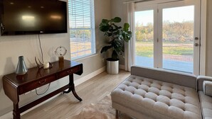 Una sala de estar con una televisión de pantalla plana, una consola de madera, un sofá color beige, una planta grande en maceta y un ventanal corredero que da a un espacio exterior.