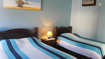 A room with two beds, each with striped bedding, a nightstand with a lamp, a painting on the wall, and an air conditioning unit.