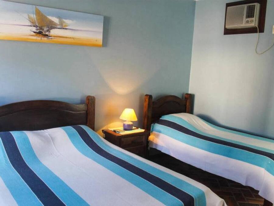 A room with two beds, each with striped bedding, a nightstand with a lamp, a painting on the wall, and an air conditioning unit.
