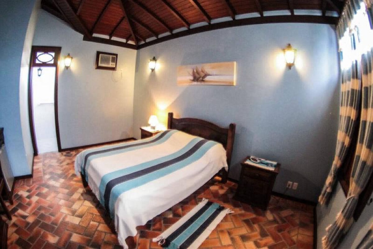A bedroom with a bed, a nightstand, a lamp, a painting on the wall, and a herringbone-patterned floor.