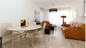 Ein modernes Wohnzimmer mit einem runden Esstisch, beigen Stühlen, einem braunen Ledersofa, einer schwarzen Stehlampe und einem Fernseher.