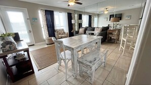 A spacious living room with a dining area, large windows with curtains, ceiling fans, and a kitchen area with white furniture.