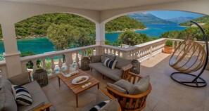 A terrace with a sofa set, a dining table, and a hanging chair, overlooking a turquoise sea and green hills. - Beachfront Villa Feodora Grande with Pool (Žrnovo)
