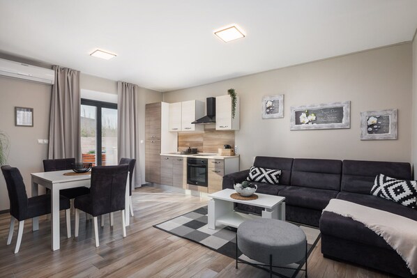 Una sala de estar moderna con un sofá negro, una mesa de comedor con sillas, una zona de cocina y un ventanal con cortinas.