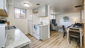 A compact kitchen with white cabinets, a refrigerator, a stove, and a sink. There is a dining area with a wooden table and chairs. - #HabitueHomes - Red Ivy #4 - 2 Bed + Pullout Sofa + Dog Friendly (Boise)