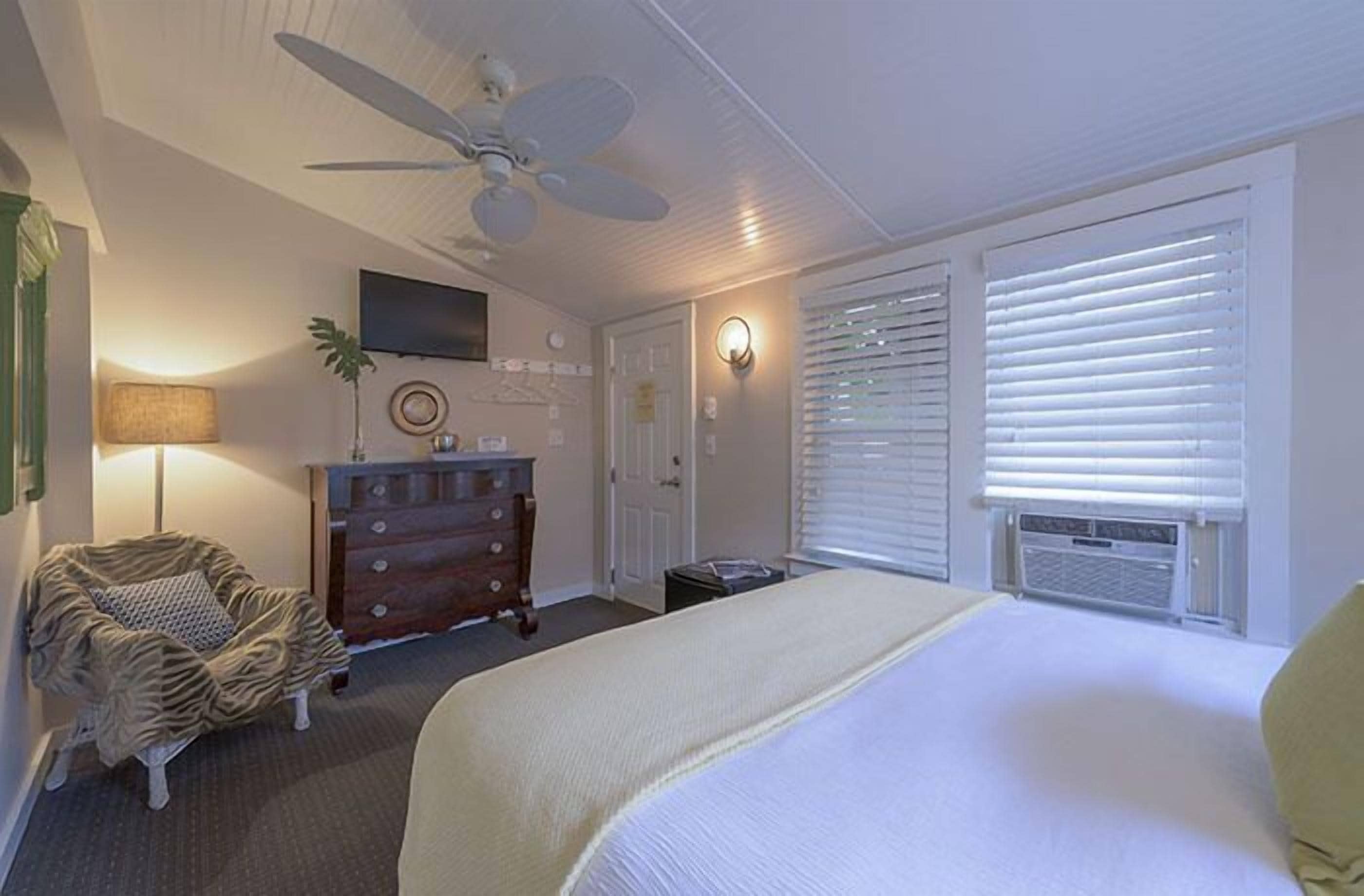 A bedroom with a bed, a dresser, a chair, a ceiling fan, and a television on the wall.