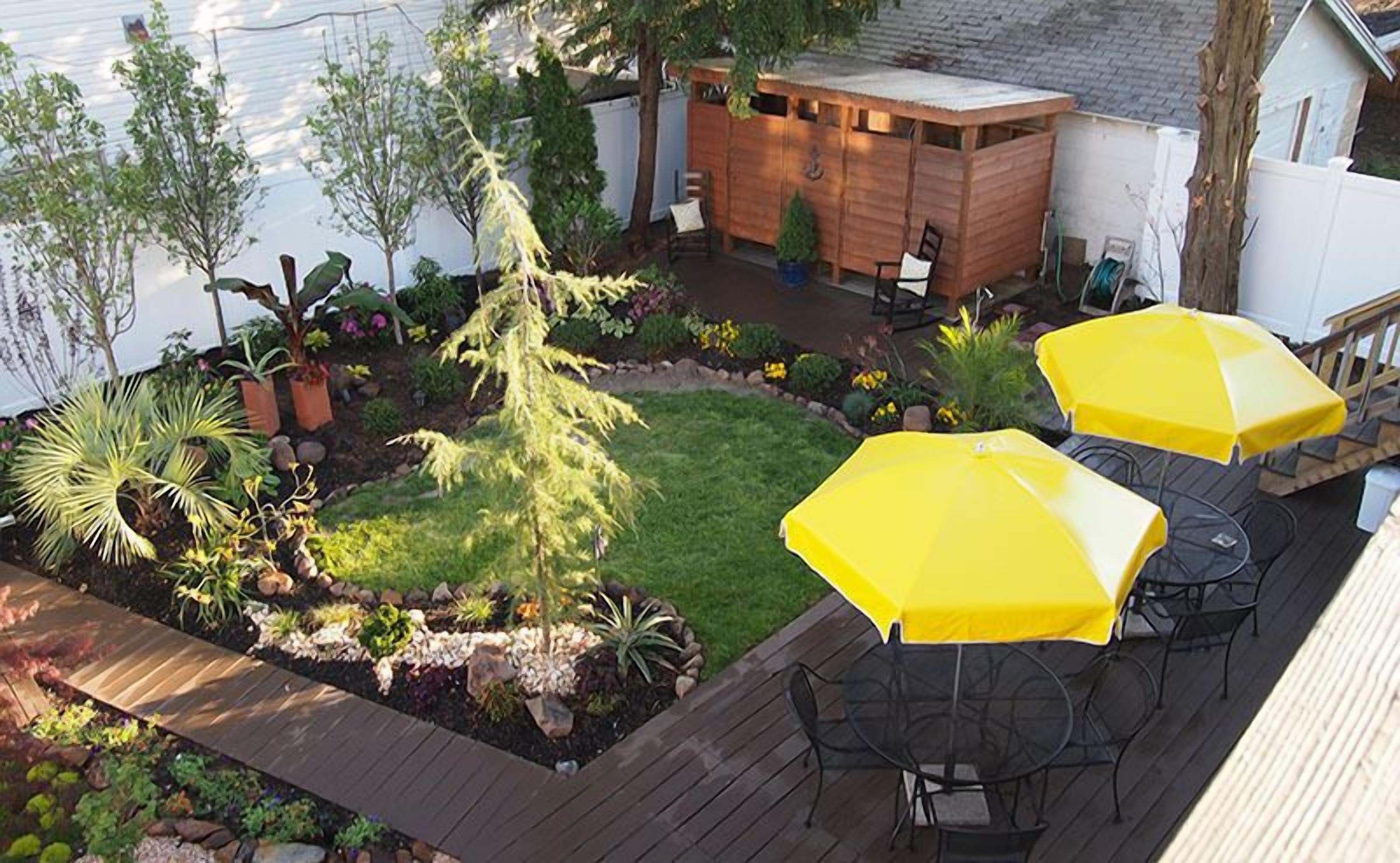 Een achtertuin met een houten terras, twee gele parasols en verschillende planten en bloemen.