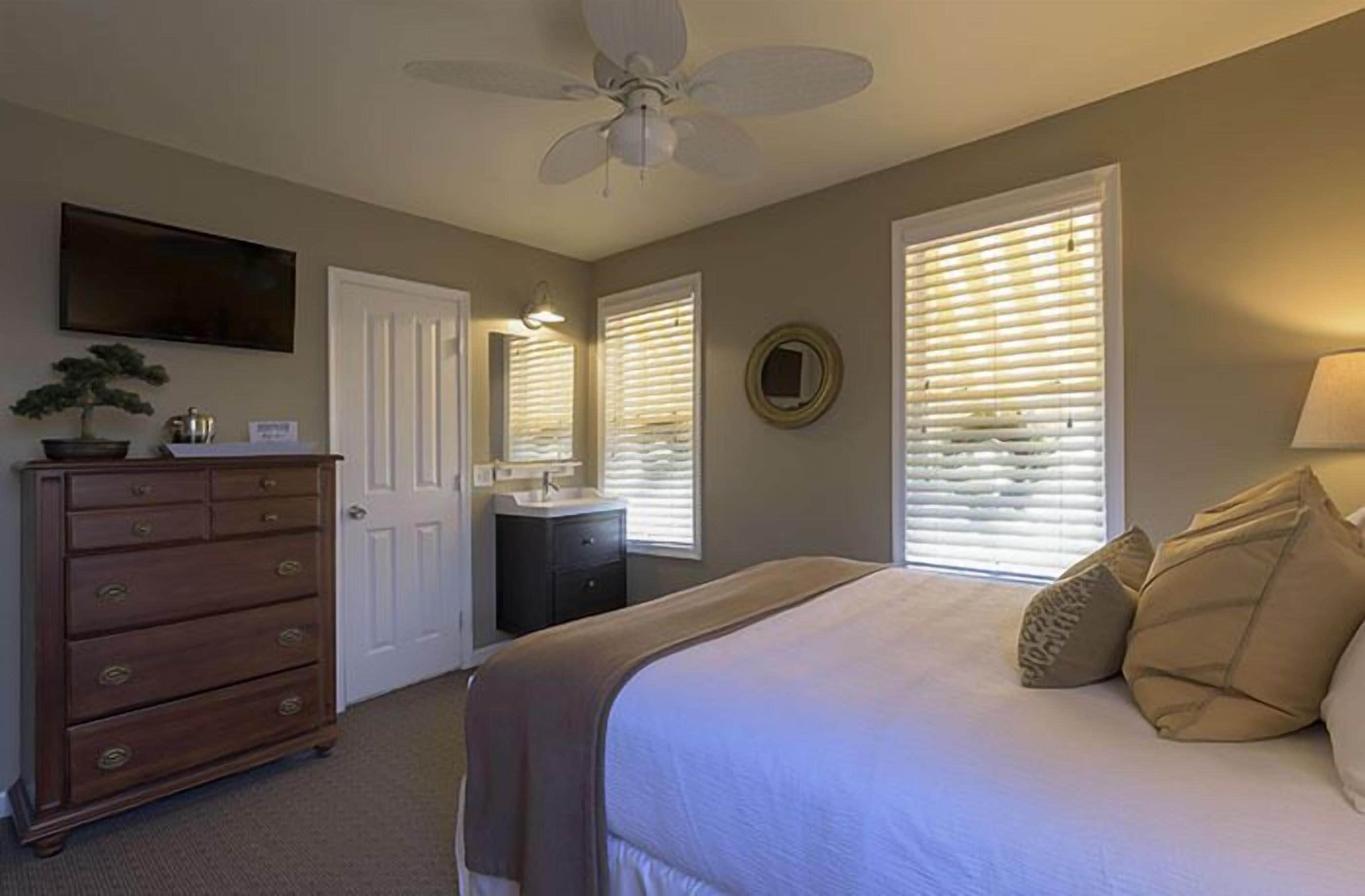 A bedroom with a bed, dresser, ceiling fan, and a TV mounted on the wall.