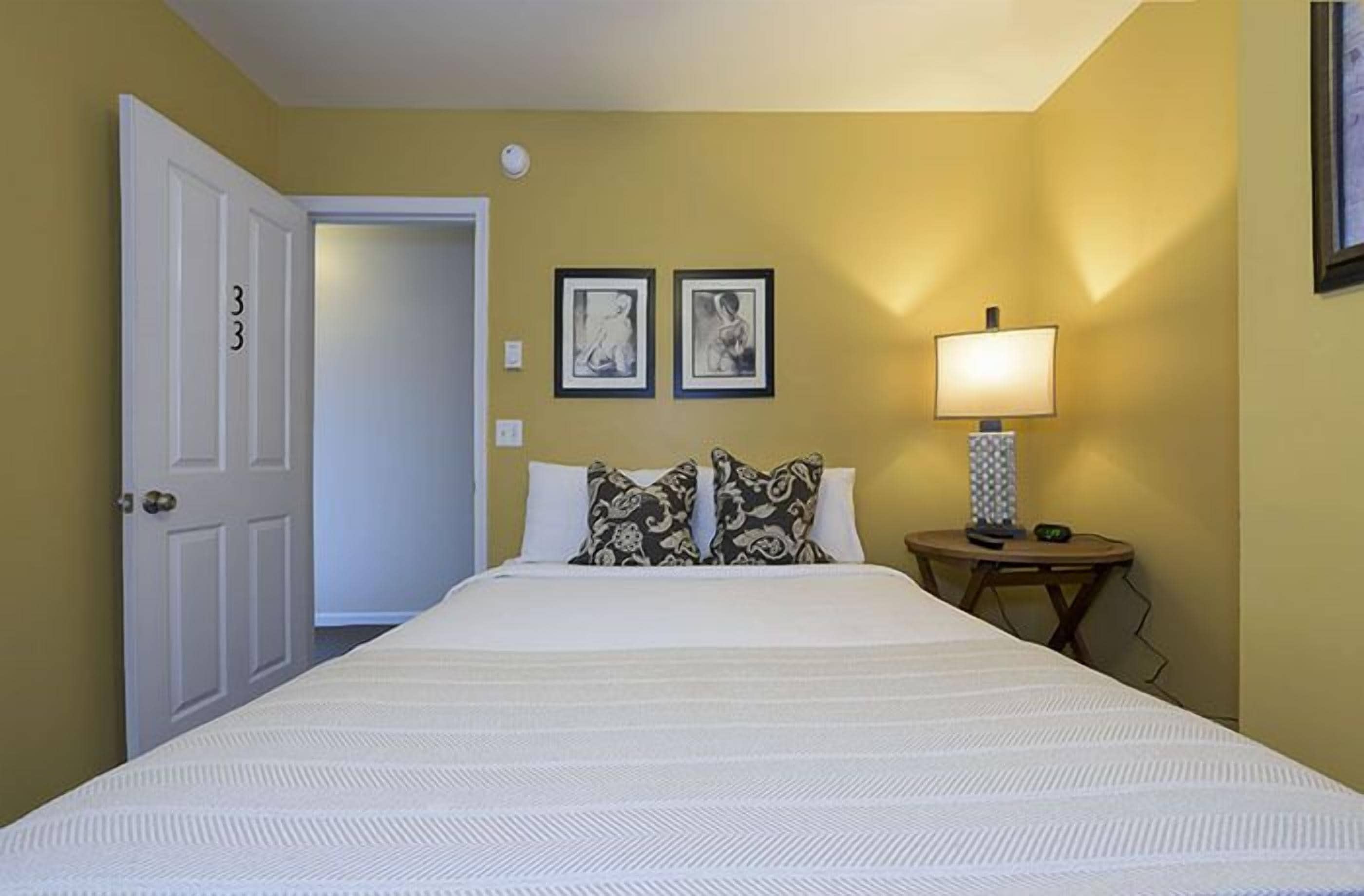A hotel room with a large bed, two framed pictures on the wall, a bedside lamp, and a door with the number 33.