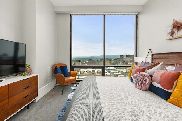 Une chambre moderne dotée d’une grande fenêtre, d’un téléviseur à écran plat, d’une commode en bois, d’une chaise et d’un lit agrémenté de coussins colorés.