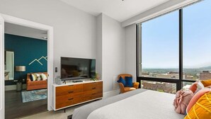 A modern bedroom with a large bed, a TV, a dresser, and a view of the city.