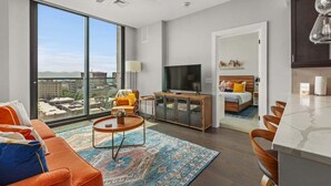 A modern hotel room with a large window, a flat-screen TV, a bed, a dining area, and a ceiling fan.
