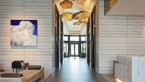 A modern hotel lobby with a long reception desk, a seating area with a sofa and armchairs, a large abstract painting, and a ceiling with leaf-like light fixtures.