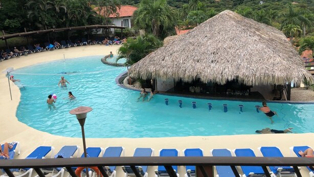 A pool area with lounge chairs, a thatched-roof hut, and a view of a hillside with buildings. - Beautiful Costa Rican Villa in Gated Beach Resort (Playa Hermosa)