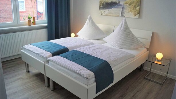 Un letto matrimoniale con biancheria da letto bianca e azzurra, un comodino con una lampada, una finestra vista su un edificio e un quadro appeso al muro.