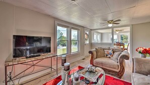 A spacious living room with a large flat-screen TV, comfortable seating, and large windows offering a view of the outdoors. - Porch, Fire Pit & Grill: Blue Ridge Mtn Cottage! (Hudson)