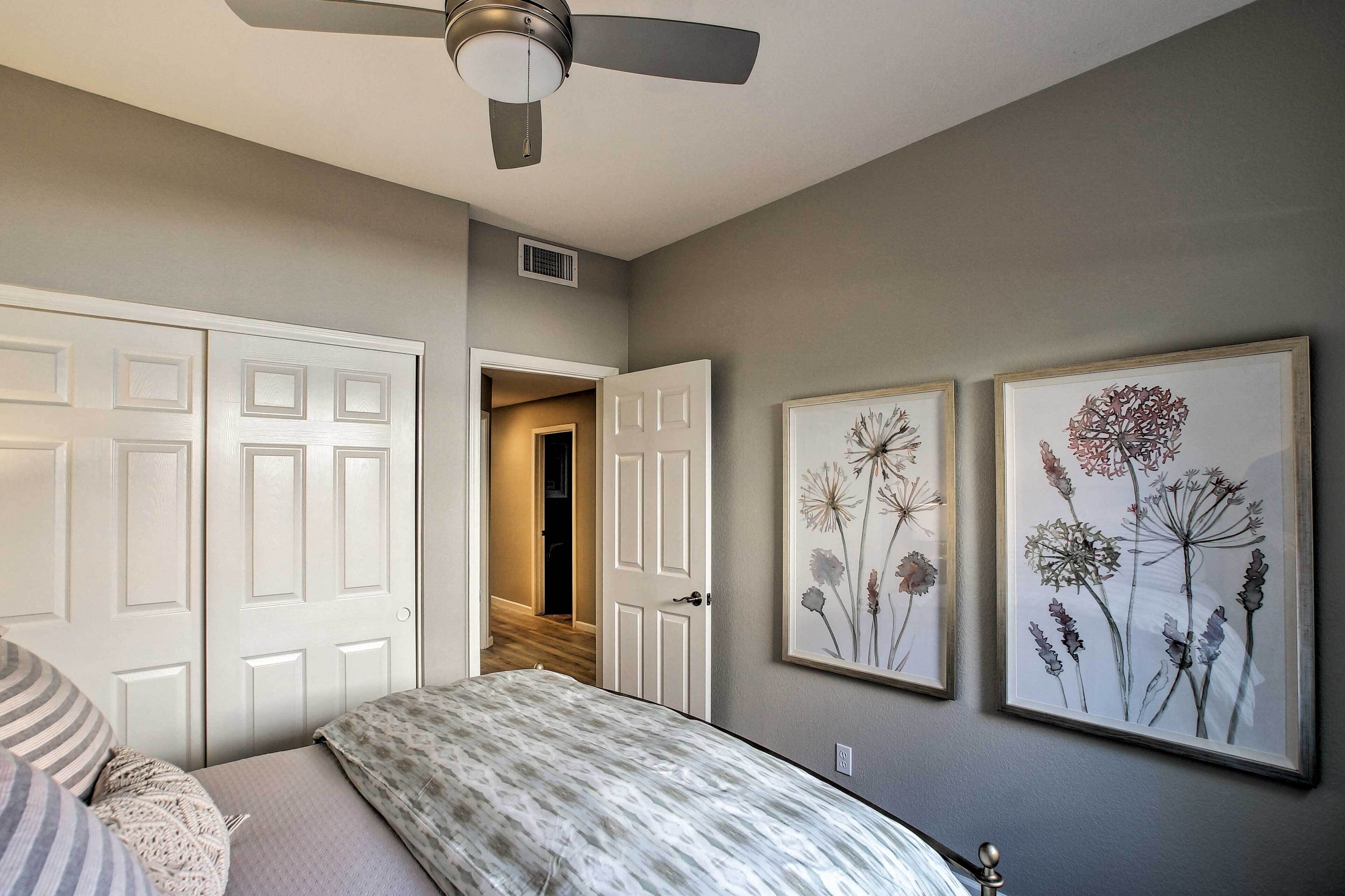 A bedroom with a bed, a ceiling fan, two framed floral artworks, and a door leading to another room.